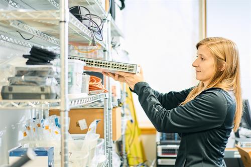 Organizing the network: Technician Kireina placing a switch among shelves of tech gear and cables in a sunlit workspace.