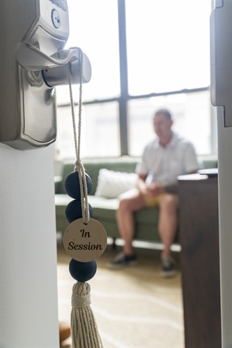 An "In Session" sign hangs outside a bright Chicago therapy office with a person seated in the background.