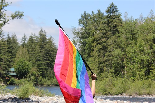 Close up photo of Gilbert Baker Pride flag flying with green trees in the background