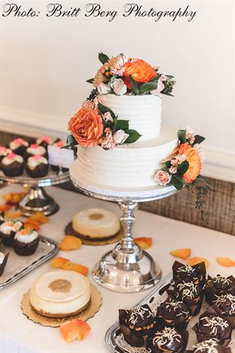 Sweets table with two-tier cutting cake, cheesecakes, and gourmet cupcakes accented by orange flowers