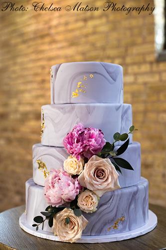 Four-tier cake with a purple marbled fondant overlay, edible gold leaf detailing, and bold floral