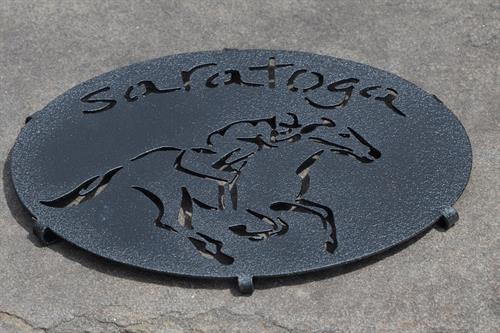 Saratoga text arched over a jockey riding a horse on a metal trivet; powdered paint in black