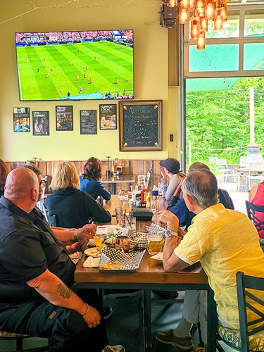 Indoor and outdoor dining at Artisanal Brew Works in Saratoga Springs, featuring World Cup soccer viewing, forest views, great food, refreshing beverages, and free parking.