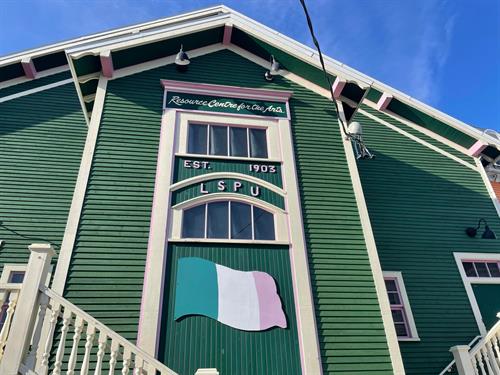 A photo of the front of the Hall, a green building with pink and white trim. Eight small windows in two lines of four and a green, white, and pink NL republic flag.