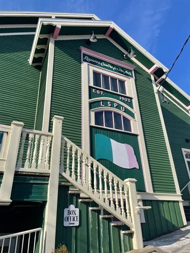 A vertical photo of the Hall, a green building with pink and white trim. Eight small windows in two lines of four and a green, white, and pink NL republic flag.