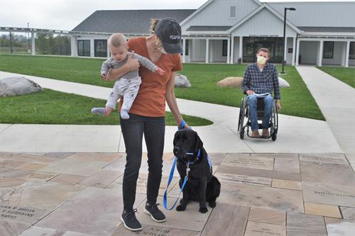 A Canine Companions client working hard with her service dog, holding her baby, while her husband watches from afar. 
