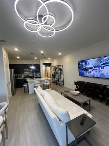  Living Combo with Gold Accents This cozy yet polished layout features a granite-topped island with plush bar stools, pendant lights suspended by gold chains, and a welcoming living area anchored by a textured sofa and wood coffee table. The dark blue cabinetry and light tile flooring create a crisp contrast that feels both fresh and grounded.