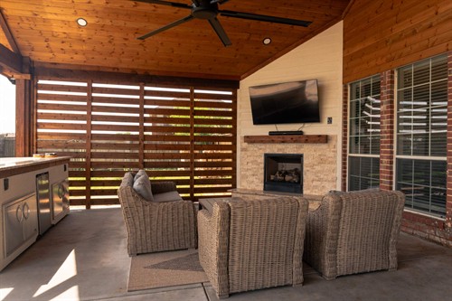 Outdoor living space with a gas fireplace, slat wall for a combination of privacy & shade. TV mounted above a mantel, 8' wide fan above, and an outdoor kitchen over looking a pool.