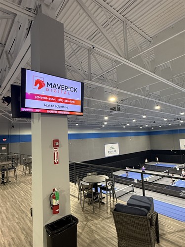 An elevated view of an indoor pickleball facility with a Maverick Digital display screen mounted on a pillar overlooking the courts.