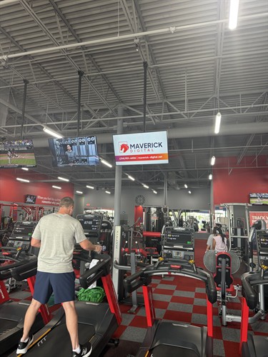 A person working out on a treadmill with an overhead TV screen displaying the Maverick Digital logo and contact information.