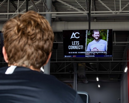 A view from behind a gym-goer looking up at a Maverick Digital indoor billboard showcasing a local partner's advertisement.
