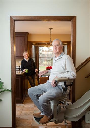 Gallery Image man-riding-curved-stairlift-with-wife-in-background-holding-coffee-mug.jpg