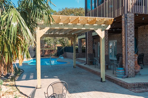 A handcrafted wooden pergola stands on a paved poolside patio, featuring heavy timber construction and decorative end cuts on the rafters. The structure is anchored with secure black metal base mounts onto a stone paver floor, situated next to a brick home with a multi-level deck. Lush palm leaves frame the foreground, while a clear blue swimming pool and mature trees provide a relaxing backdrop.