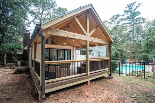 A spacious, vaulted-roof screened porch addition features natural wood timber framing and exposed trusses. The interior includes a dark composite deck floor, a black ceiling fan, and recessed lighting. A brown railing with black balusters provides a clear view of a backyard swimming pool and surrounding mature trees.