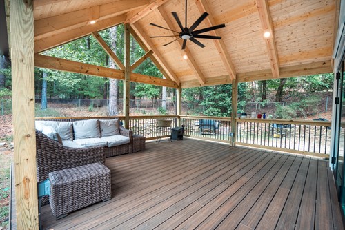 Interior of a custom screened-in porch featuring a high vaulted ceiling with light-colored wood planks and a large black ceiling fan. The space is furnished with a grey wicker sectional sofa on a dark composite deck floor. Recessed lighting and black metal railings complete the modern outdoor living area.