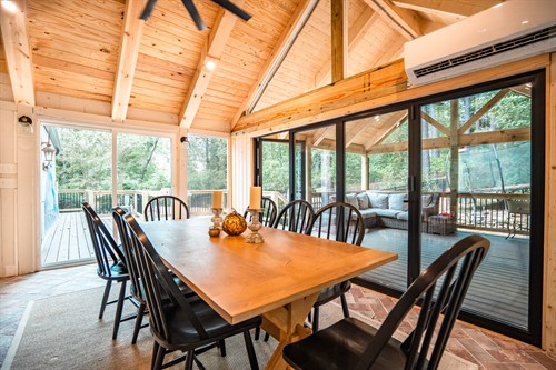 Interior dining area featuring a wooden table with black chairs situated on a light-colored rug. Large black-framed sliding glass doors offer a clear view into an adjacent screened-in porch with a wicker sectional. The space includes a vaulted wood ceiling, recessed lighting, and a wall-mounted mini-split AC unit.