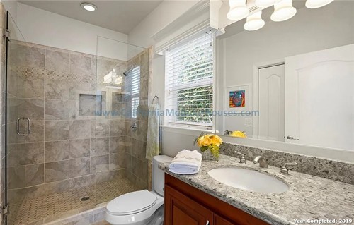 Bathroom with a walk-in shower enclosed with glass, a toilet, a granite countertop with a sink, window with white blinds, and a white door. California Modular Houses. Modular home construction California.  Prefab house.  Oxnard, Ventura County, CA.  