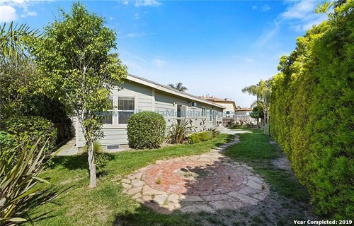 View of a backyard with a green house, lush green trees and bushes, a stone pathway, and a green hedge on a sunny day.  California Modular Houses. Modular home construction California.  Prefab house.  Oxnard, Ventura County, CA.  