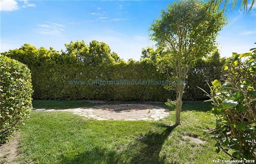 A backyard with a grassy lawn, small trees, and a stone patio area enclosed by a hedge fence under a clear blue sky.  California Modular Houses. Pre fab home.  Modular homes for California.  Casas prefabricadas.  Southern California.  