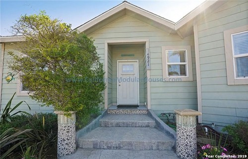 Front entrance of a light green house with white trim, featuring a white front door, two windows, a small porch with concrete steps, and decorative mosaic columns on either side of the steps.   California Modular Houses. Modular home construction California.  Prefab house.  Oxnard, Ventura County, CA.  