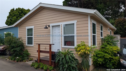 A beige single-story house with white trim and a small front porch with a wooden railing. The house has four windows, with blinds visible through the glass. There are green bushes and plants around the house, and part of a driveway with vehicles on either side. A black outdoor wall light is mounted next to the front door.