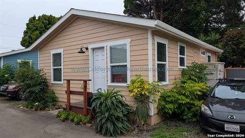 A small beige house with white trim, green plants along the front, and a black Mazda car parked to the right. A white door with a small porch and wooden steps is visible. The house has four windows and an outdoor light fixture.