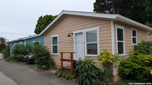 A single-story house with beige and blue siding, white trim, and multiple windows, surrounded by greenery and plants, with a small wooden porch and a parked car nearby.