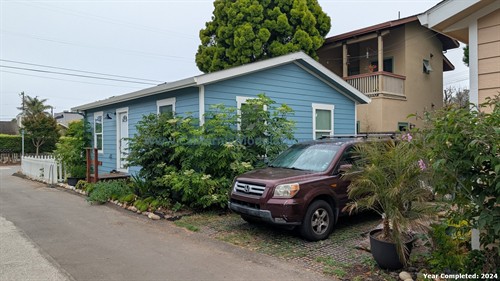 A small blue house with white trim, surrounded by greenery and flowers, with a maroon Honda Pilot parked in front, and two neighboring buildings visible in the background on a cloudy day.