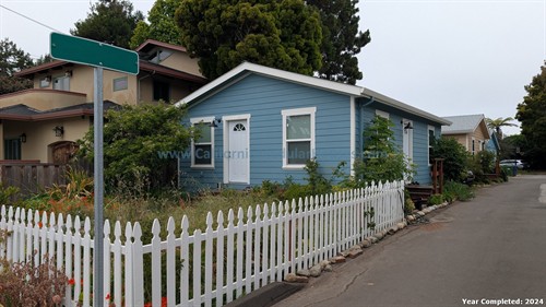 A blue house with a white picket fence along a street. The house has white trim and a small front porch. Surrounding garden with bushes and small trees. Nearby houses are visible in the background, and a large tree extends over the roof. An empty street sign pole is in the foreground.