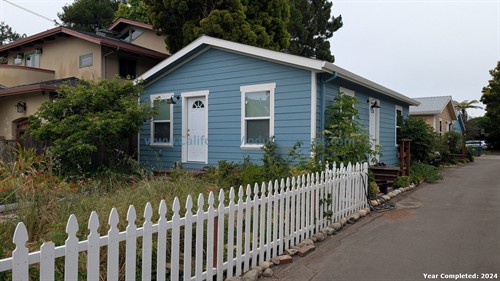 A small blue house with white trim and a white picket fence, surrounded by greenery, in an urban neighborhood.
