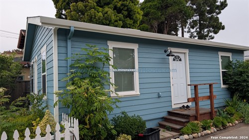 Small blue house with white trim, three windows, wooden steps, black outdoor light, and surrounding greenery.