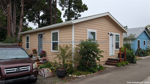 A beige single-story house with white trim and a small wooden staircase at the front door, surrounded by plants and flowers, with a maroon SUV parked in front on a paved driveway. There are trees in the background and neighboring houses painted blue.