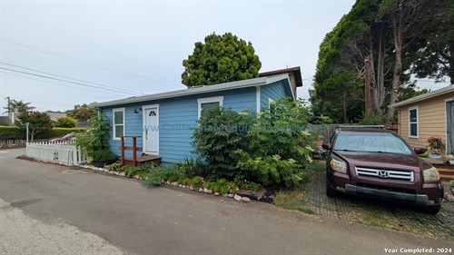 A small blue house with white trim, a white door with a small window, and gray roof. A small wooden ramp leads to the door. There are plants along the house with a white picket fence on the left. On the right, there is a brown Honda SUV parked on a brick driveway. Tall trees and other homes are visible in the background.