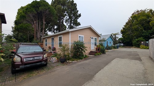 A residential area with a beige house with white trim, surrounded by plants and flowers, with a maroon SUV parked in front. There are tall trees behind the house and a blue house in the background, along with parked cars and a driveway.
