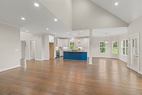 Spacious open-concept living area with soaring vaulted ceilings, rich hardwood floors, and bright white kitchen featuring a bold blue island.