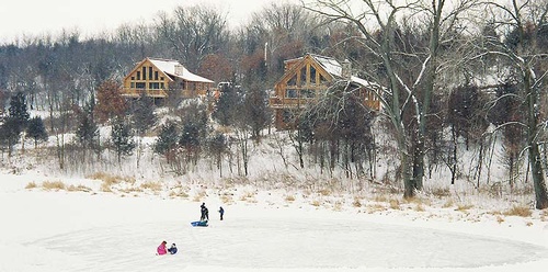 view of two log cabins during the winter with a family sledding