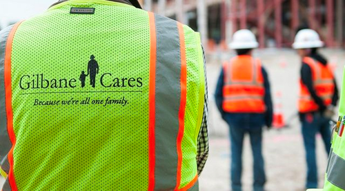 Gilbane Building Company team standing outdoors onsite. Person facing away, wearing a Ã¢??Gilbane CaresÃ¢?ï¿½ neon vest. 